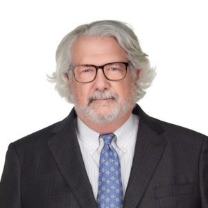 An older man with gray hair and a beard is wearing glasses, a dark suit, a striped shirt, and a blue patterned tie against a plain white background.