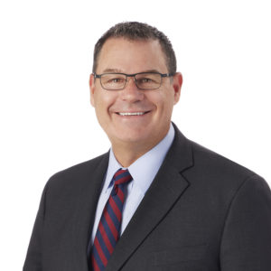 A person in formal attire, wearing glasses and a suit with a striped tie, smiles warmly against a white background.