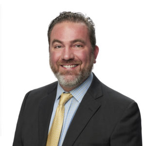 A bearded man wearing a suit and tie smiles in front of a white background.