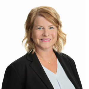 A person with blond hair is smiling and wearing a dark blazer over a light blue top.