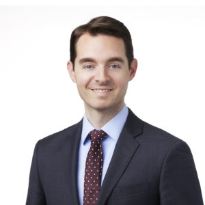 A smiling man in a suit and tie stands against a plain white background.
