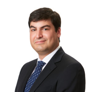 A person in a suit and tie stands against a white background, looking directly at the camera.