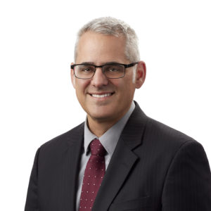 A man in a dark suit, red tie, and glasses is smiling against a white background.