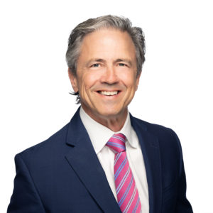 A man with gray hair is smiling while wearing a navy blue suit, white shirt, and pink striped tie against a plain white background.