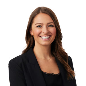 A person with long brown hair, wearing a black blazer and black top, is smiling against a white background.