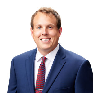 A person is smiling while wearing a blue suit, white shirt, and red-striped tie against a white background.