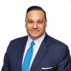 A person dressed in a navy blue suit and blue tie smiles against a white background.