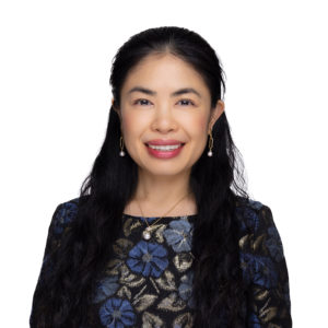 A woman with long, dark hair smiles while wearing a floral-patterned top and pearl earrings against a white background.