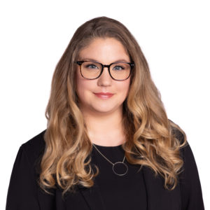 A person with long, wavy blonde hair and glasses is wearing a black top and a circular necklace.