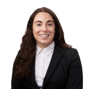 A person with long, wavy hair is smiling and wearing a black blazer over a white button-up shirt against a plain white background.