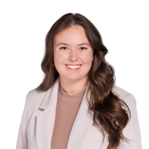 A woman with long, wavy brown hair is smiling while wearing a light-colored blazer over a beige top.