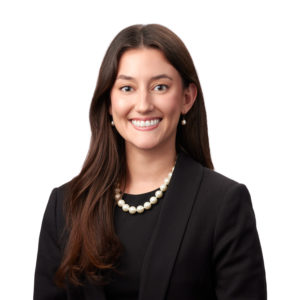 A person with long brown hair is wearing a dark blazer, a black top, and a pearl necklace, smiling against a white background.