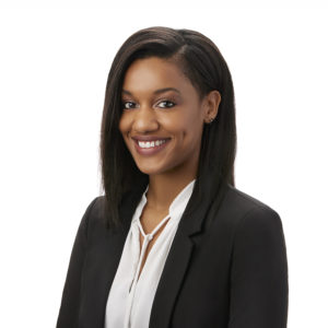 A person with shoulder-length hair is smiling, wearing a black blazer over a white blouse against a white background.