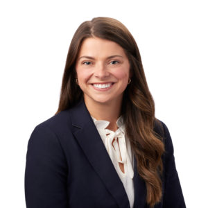A person with long brown hair is smiling, wearing a dark blazer and a white blouse, posed in front of a plain white background.