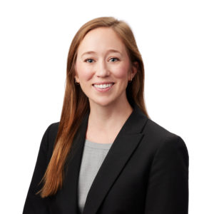 A person with long blonde hair is wearing a black blazer over a light gray shirt and smiling against a white background.