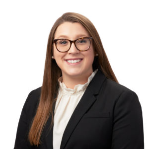 A person with long brown hair and glasses is wearing a black blazer over a white blouse against a white background.
