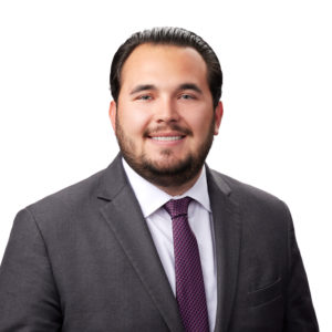 A man is dressed in a dark suit with a white shirt and purple tie, smiling against a plain white background.