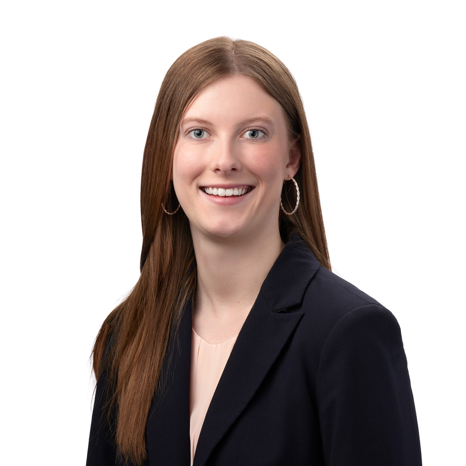 A smiling woman with long brown hair is wearing a dark blazer and hoop earrings against a white background.