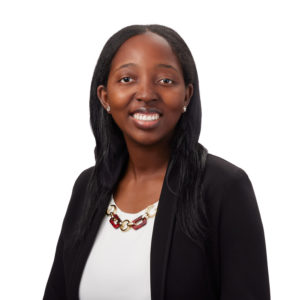 A person with long black hair is smiling and wearing a black blazer, white shirt, and a statement necklace with red and gold accents.