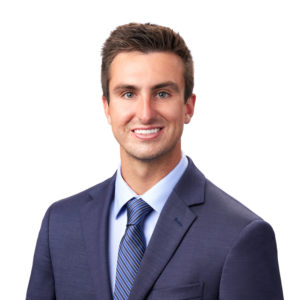 A man wearing a dark blue suit, light blue dress shirt, and striped tie smiles against a plain white background.