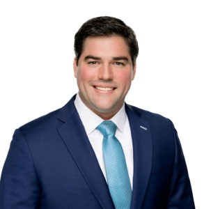 A man in a blue suit and light blue tie is smiling against a white background.