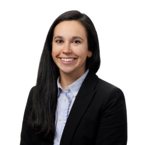 A person with long dark hair is smiling while wearing a black blazer over a light blue button-up shirt against a white background.