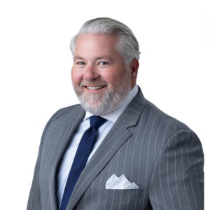 A smiling man with gray hair and a beard is wearing a gray pinstripe suit, white shirt, and blue tie against a white background.