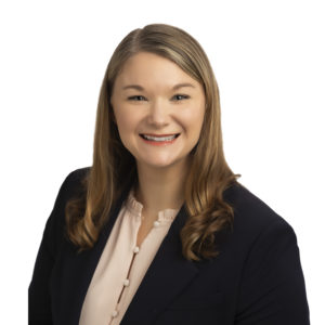 A smiling individual with long, light brown hair wearing a navy blazer and a light pink blouse posing in front of a white background.