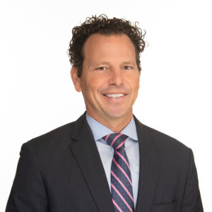 A smiling person with curly hair is wearing a dark suit and a striped tie against a plain white background.