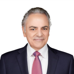 A person with grey hair, wearing a suit, white shirt, and red tie, smiles against a plain white background.