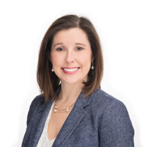 A woman with shoulder-length brown hair is smiling while wearing a grey blazer, white blouse, and pearl earrings.