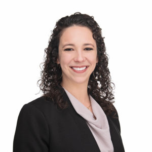 A smiling individual with curly hair, dressed in a black suit jacket and light-colored blouse, poses against a white background.