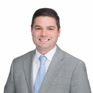 A person in a gray suit and light blue tie smiles against a plain white background.