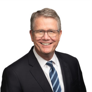 An older man in a suit and striped tie is smiling against a plain white background.