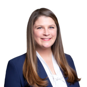 A person with long, straight hair is wearing a navy blazer over a white blouse, smiling against a plain white background.