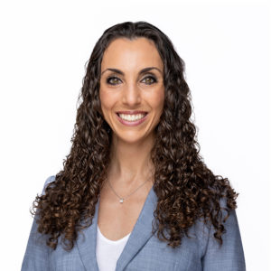A woman with long curly hair wearing a blue blazer smiles against a white background.