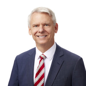 A smiling individual with white hair is wearing a dark suit, white shirt, and a red and white striped tie against a plain white background.