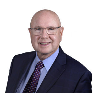 A person with glasses is smiling and wearing a suit with a blue shirt and patterned tie against a white background.