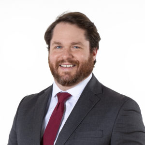 A man with a beard is wearing a dark gray suit, white shirt, and red tie, smiling at the camera against a plain white background.