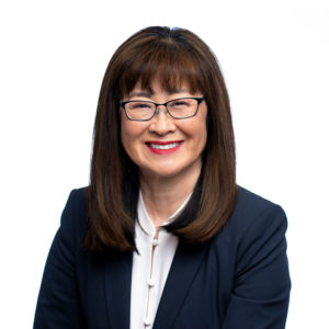 A smiling person with glasses, wearing a dark blazer over a white blouse, is posed against a plain white background.