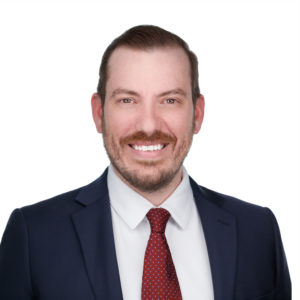 A man wearing a dark suit, white shirt, and red tie is smiling against a plain white background.