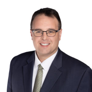 A man with glasses is wearing a dark suit and a patterned tie, smiling against a white background.
