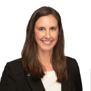 A smiling woman with long brown hair is wearing a black blazer over a white top against a plain white background.