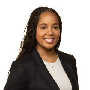 A person is smiling while wearing a black blazer and white shirt with a cross necklace.